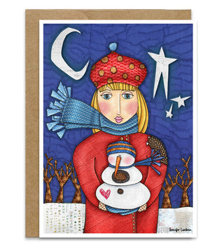 A winter and Christmas themed greeting card featuring a stylized illustration of a girl in a red coat and hat holding a snowman, with a blue night sky, crescent moon, stars, and bare trees. The card is shown with a brown kraft envelope behind it on a white background. Artwork by Jennifer Lambein.