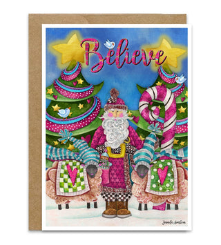 A Christmas greeting card with a kraft envelope, featuring a whimsical illustration of Santa Claus wearing a pink suit, sheep, candy canes, and trees under the word 'Believe' in pink script. Artwork by Jennifer Lambein.