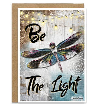 Greeting card with dragonfly illustration and 'Be The Light' text on a wooden background.