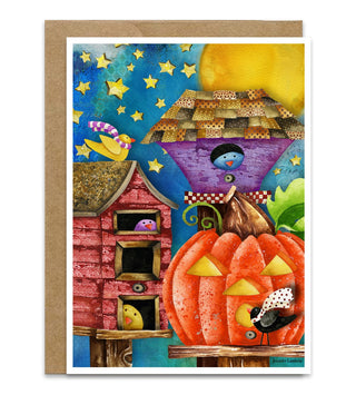 A greeting card with a whimsical Halloween illustration, shown with a brown kraft envelope behind it on a white background. The artwork features a large orange jack-o'-lantern, a red house, a purple birdhouse, yellow stars, and a blue night sky. Artwork by Jennifer Lambein. 
