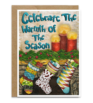 A square greeting card with a kraft envelope, featuring a festive illustration of colorful Christmas stockings, lit candles, and green garland beneath the text 'Celebrate The Warmth Of The Season' in teal lettering. Artwork by Jennifer Lambein. 