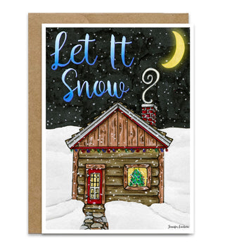 A greeting card featuring a winter illustration of a log cabin in the snow under a black night sky with a yellow crescent moon and the text 'Let It Snow?'. The card is shown with a brown kraft envelope behind it. Artwork by Jennifer Lambein. 