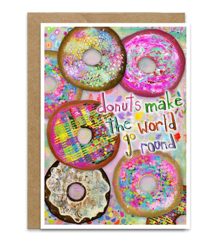 A greeting card with a kraft envelope, featuring a colorful illustration of donuts and the text 'donuts make the world go round' on a white background. Artwork by Jennifer Lambein. 