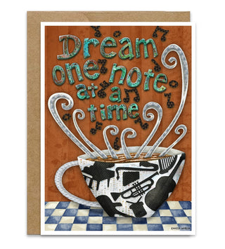 A greeting card with a kraft envelope, featuring an illustration of a coffee cup with musical instrument silhouettes on it, with musical notes surrounding it and the text 'Dream one note at a time' on a warm brown background. Artwork by Jennifer Lambein. 