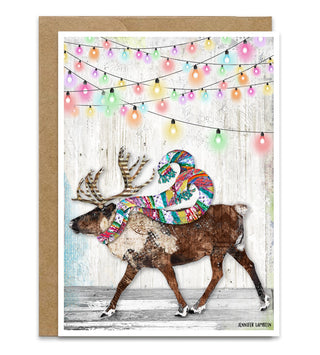 A greeting card featuring an artistic illustration of a brown reindeer wearing a colorful patterned scarf, walking under pastel-colored string lights against a whitewashed wood background. The card is shown with a brown kraft envelope behind it on a white studio background. Artwork by Jennifer Lambein. 