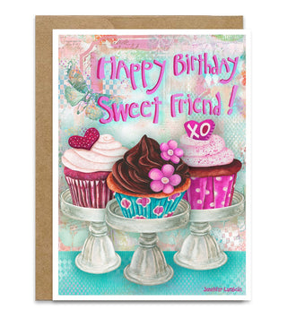 A birthday greeting card featuring three decorated cupcakes on glass stands with the text 'Happy Birthday Sweet Friend!' in pink script, shown with a brown kraft envelope behind it on a white background. Artwork by Jennifer Lambein. 