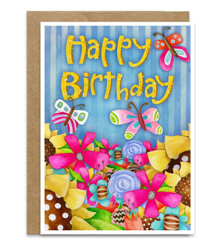 A colorful 'Happy Birthday' greeting card with a brown kraft envelope, featuring glittery gold lettering, butterflies, and a vibrant floral and sunflowers collage on a white background. Artwork by Jennifer Lambein. 