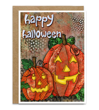 A greeting card with a kraft paper envelope behind it, featuring two watercolor carved jack-o'-lantern pumpkins and the words 'happy halloween' in white lettering on a textured autumn background. Artwork by Jennifer Lambein.