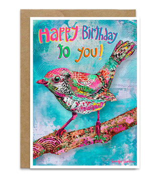 A colorful birthday card featuring a vibrant, patterned bird perched on a branch with the text 'Happy Birthday to you!' in playful multicolor lettering, shown with a brown kraft envelope behind it on a white background. Artwork by Jennifer Lambein. 