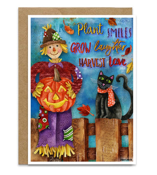 An autumn and Halloween greeting card with a kraft paper envelope, featuring a colorful Halloween illustration of a smiling scarecrow holding a jack-o'-lantern and a black cat wearing a red scarf. The card reads, 'Plant smiles GROW laughter HARVEST love' with autumn leaves scattered around. Artwork by Jennifer Lambein. 