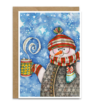 A Christmas greeting card featuring a whimsical watercolor-style snowman wearing a red and green striped beanie and scarf, holding a mug with steam, set against a blue starry winter background. The card is shown with a brown kraft envelope behind it on a white studio background. Artwork by Jennifer Lambein. 