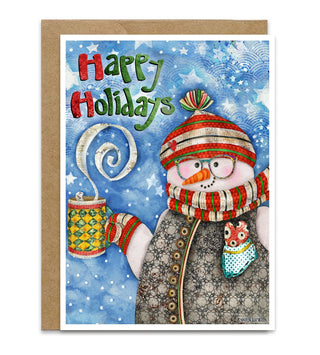 A Christmas holiday greeting card with a kraft envelope behind it, featuring a whimsical snowman illustration wearing a red-and-white striped hat and scarf, holding a mug, with the words “Happy Holidays” in red and green lettering on a blue snowy background. Artwork by Jennifer Lambein. 