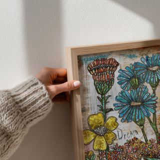 Hand holding a wooden-framed artwork with colorful floral designs on a white wall.