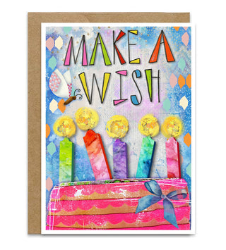 A colorful birthday greeting card with a kraft envelope, featuring the text 'MAKE A WISH' above illustrated candles and a pink cake on a light blue patterned background. Artwork by Jennifer Lambein. 