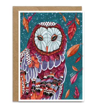 A fall greeting card featuring a detailed, colorful illustration of an owl with patterned feathers on a teal starry background, with scattered autumn leaves around it. A kraft paper envelope is placed behind the card on a white background. Artwork by Jennifer Lambein. 