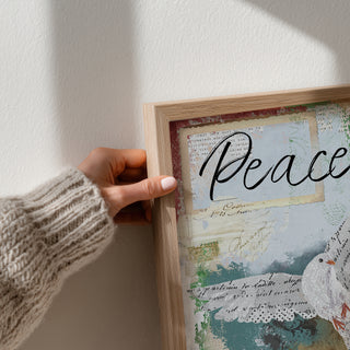 Hand holding a framed artwork with dove and the word 'Peace' on a light background