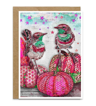 A greeting card with a kraft envelope, featuring a whimsical collage design of two birds perched on pink and green patterned pumpkins against a textured pastel sky with pink stars. Artwork by Jennifer Lambein. 