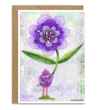 A greeting card featuring a whimsical purple flower and small bird illustration, shown with a brown kraft envelope behind it on a white background. Artwork by Jennifer Lambein. 