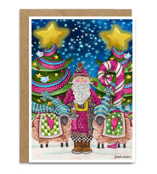 A greeting card featuring a whimsical Christmas scene with Santa in a pink suit standing in the snow with sheep, Christmas trees, and a candy cane, shown with a brown kraft envelope behind it on a starry night sky background. Artwork by Jennifer Lambein. 