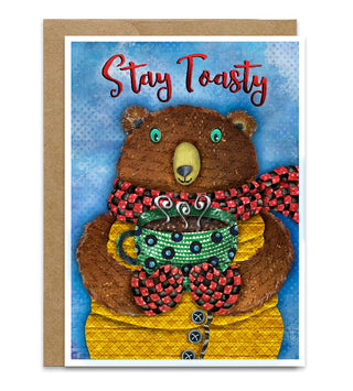A greeting card featuring a cozy illustration of a brown bear wearing a red and black checkered scarf and yellow sweater, holding a green beaded mug. The card has the words 'Stay Toasty' in red script at the top and is shown with a brown kraft envelope behind it on a white background. Artwork by Jennifer Lambein. 