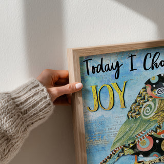 Person holding a framed artwork with 'Today I Choose Joy' text against a white wall.