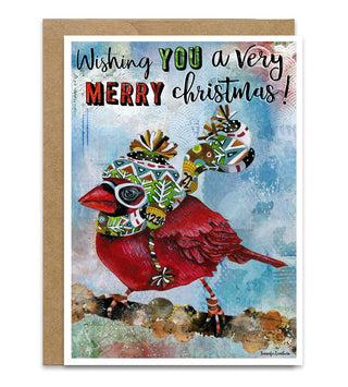 A Christmas greeting card featuring a red cardinal wearing a colorful patterned hat and scarf, with the text 'Wishing YOU a very MERRY christmas!' on a textured blue background, shown with a brown kraft envelope behind it. Artwork by Jennifer Lambein. 